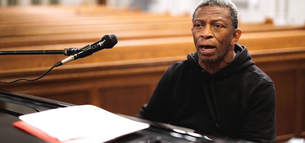 Photo: The choir director of Inner Strength Gospel Choir, Herbert Jones, leads his choir by demonstrating what to sing. His mouth is agape in the photo, mid-singing, and he wears a black hoodie.