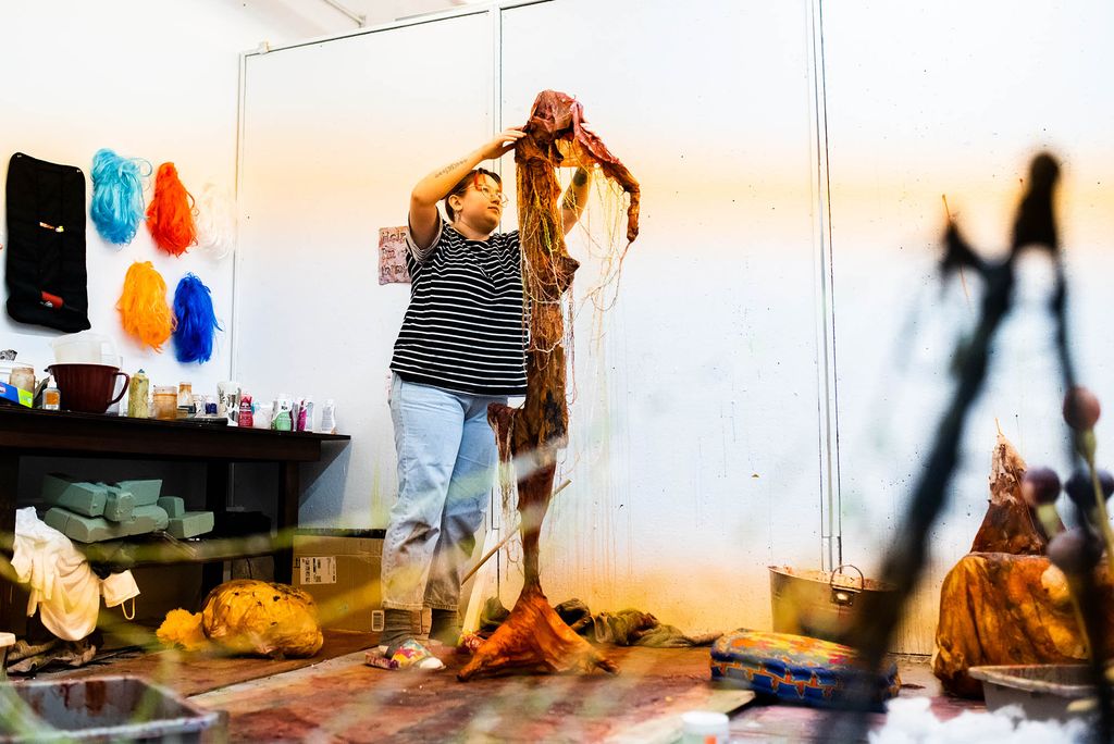 Photo: A woman with short hair adjust her sculpture for her MFA thesis. She stands in a room with no windows and the sculpture is a wire sculpture.