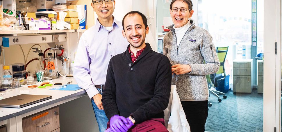 Photo of Dr. Chris Chen (left) postdoc (and former PhD student) Christos Michas(center), Dr. Alice White in their lab on March 7, 2022. Chen is Asian middle aged wearing a gray button-down shirt and glasses. Christos wears purple gloves, a dark gray sweater, and crimson pants and smiles. White wears a gray patagonia quarter zip, black pants and glasses. Behind them, a shelf of lab equipment is seen.