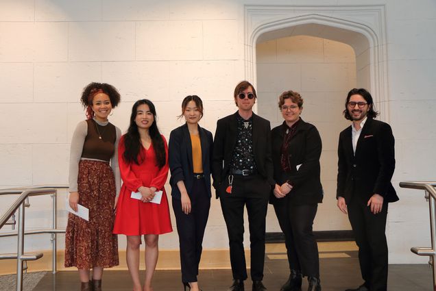 Photo: Six people stand in a line to smile at a camera for a photo. From the left, a Black woman with her hair tied up with a scarf. She wears a maroon midi skirt, a black top and gray cardigan. Next to her is a woman with long black hair and a knee-length, bright red dress. In the middle, an Western Asian woman with her hair pulled up behind her with two pieces left out to frame her face. She wears a black suit set, and an orange top. Next to her is a white male with circular glasses on and a black blazer with a patterned button up. Next, a shorter white woman with short brown hair. She wears a black suit set and a deep-colored shirt. On the end of the line, the last on the right, is a white man with shoulder length black hair. He wears a typical suit with a white button up.