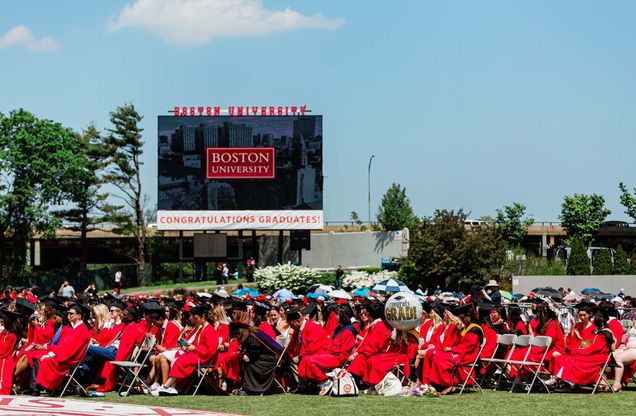 Photo: Boston University graduates sitting below the jumbotron on Nickerson field while wearing red cap and gowns
