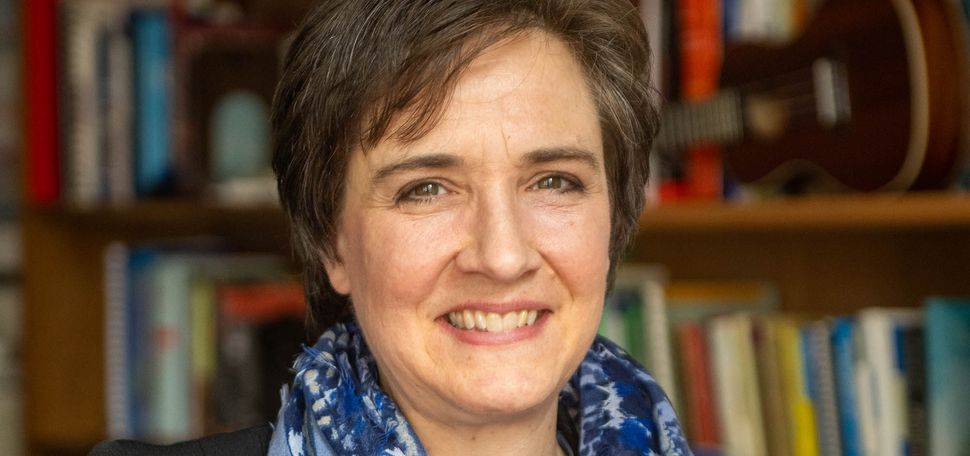 Photo: A white woman with short, brown hair looks into the camera and smiles. She wears a black cardigan and a blue-patterned infinity scarf. Behind her is a stack of books and a guitar.