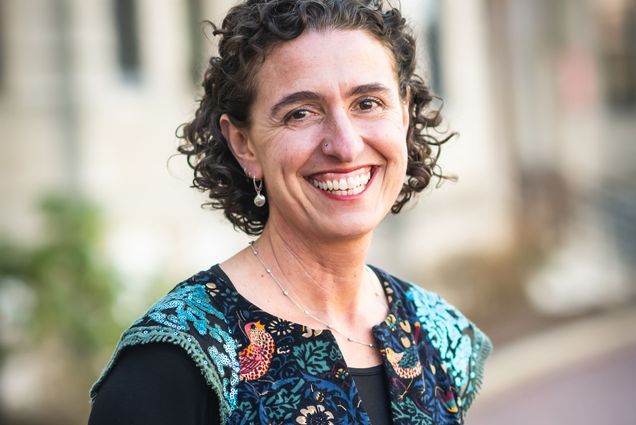 Photo: CAS professor Joanna Davidson, a white woman with short, curly brown and wearing a blue mosaic-themed blouse and black long-sleeved top smiles and poses with arms clasped behind her back. A blurry outdoor BU building can be seen behind her.