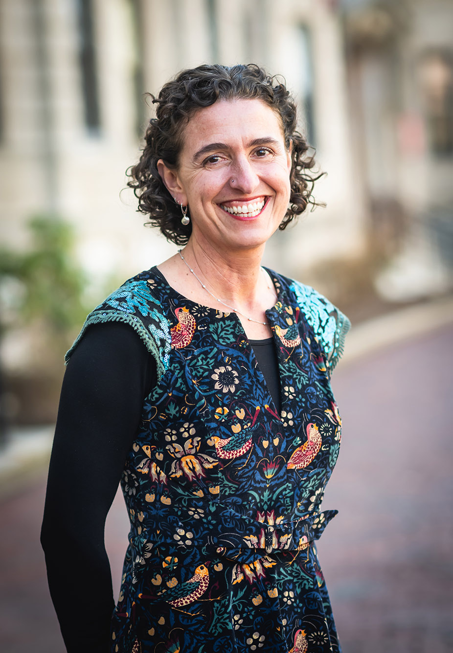 Photo: CAS professor Joanna Davidson, a white woman with short, curly brown and wearing a blue mosaic-themed blouse and black long-sleeved top smiles and poses with arms clasped behind her back. A blurry outdoor BU building can be seen behind her.