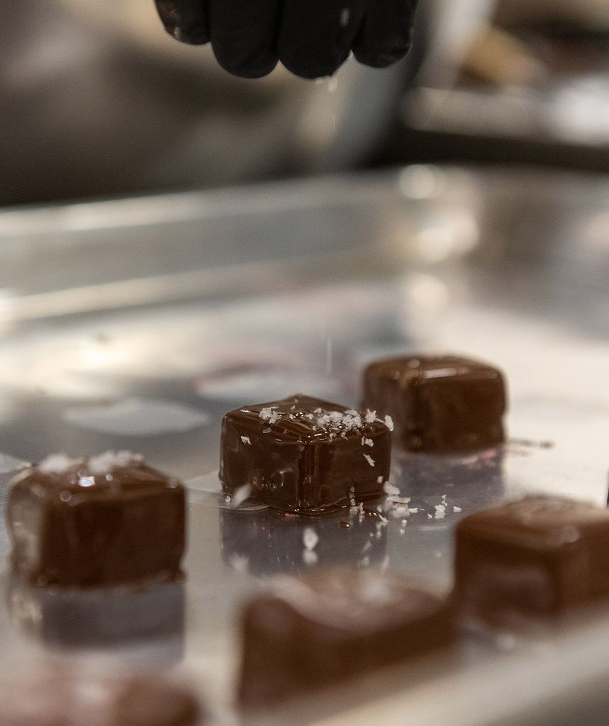 Photo: Close-up action shot of cubes of caramel dipped in tempered chocolate and being sprinkled with salt.