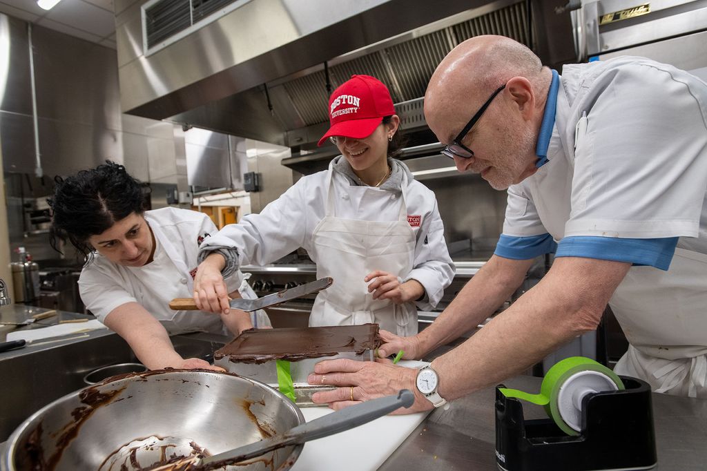 Photo: Pastry Chef Janine Sciarappa, at left, and Chef Bill Yosses, at right, help student Mara Clement (Professional Pastry Arts' 23) add ganache to her nut-free Japonaise route during class April 5. A woman wearing a white chef's outfit with sleeves rolled up (left) and an older white bald man wearing glasses and white chef's jacket (right) help a young woman (center) wearing a red Bu cap and white chef's outfit spread a layer of chocolate ganache.