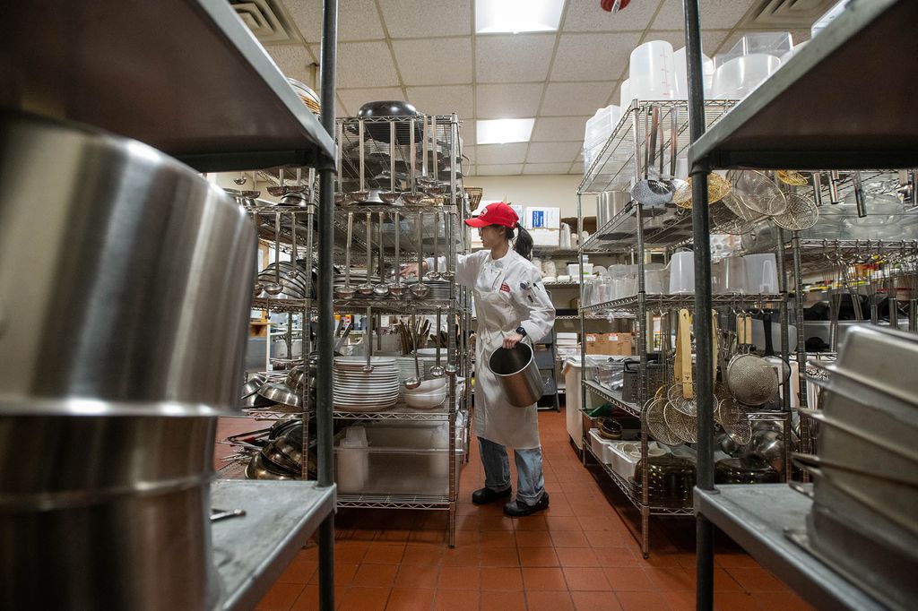 Photo: Natalie Lee, a young Asian woman wearing a red Bu cap and white chef's outfit, puts away the day’s clean bowl, pans, measuring cups, knives, pots and baking tools April 5. She places them on metal shelves in the large test kitchen.