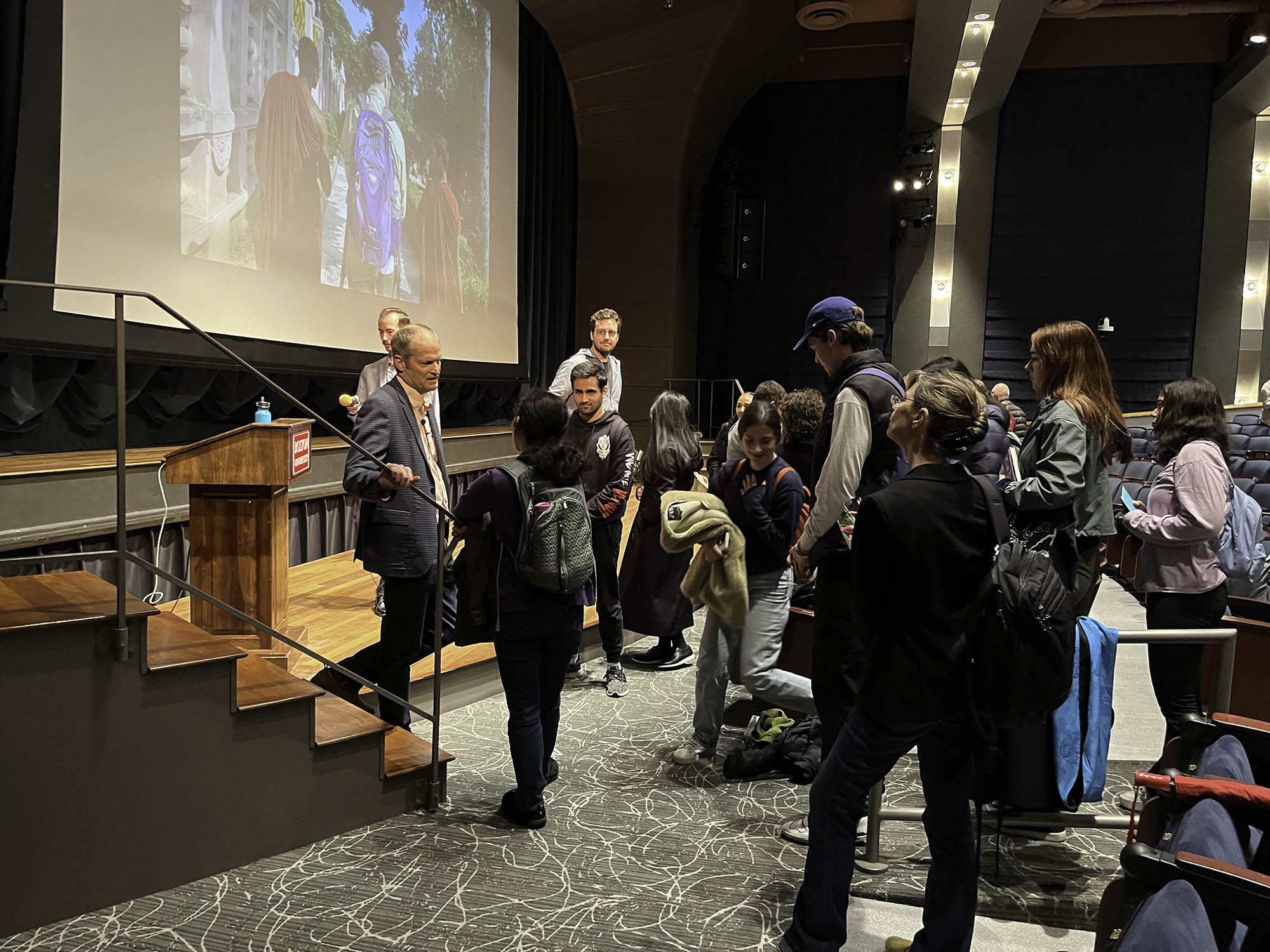 Photo: Various people and students line up in front of Prothero, a white man wearing a cream shirt, grey blazer, and slacks.