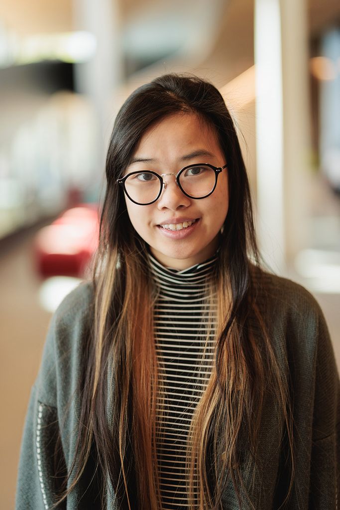 Photo: Samantha Chen, a young Asian woman with long dark brown hair with light brown highlights and wearing black glasses, a black and white striped turtleneck, and dark grey cardigan, smiles and poses for the camera.