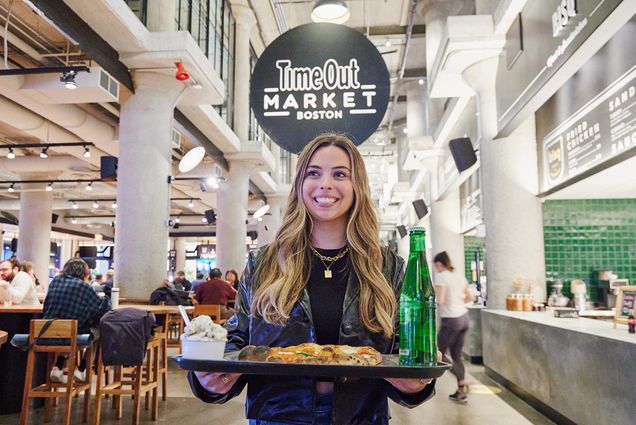 Photo: A young woman holding a tray of food stands under a sign that reads "Time Out Market"