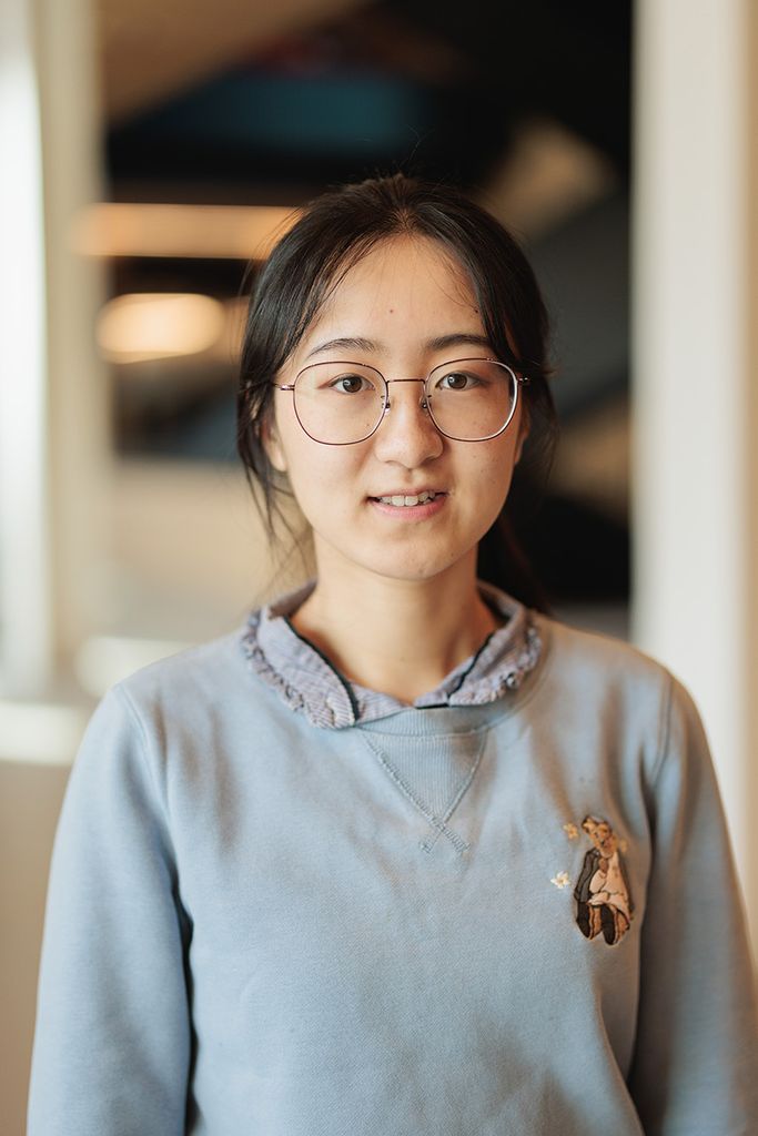 Photo: Yile Wang, a young Asian woman wearing round wireframe glasses and light blue-gray sweatshirt, smiles and poses for the camera.