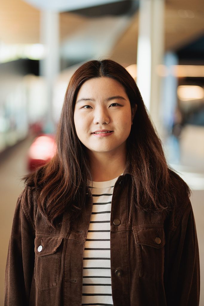 Photo: Zining Ye, a young Asian woman with long, dark brown hair and wearing a white and black striped shirt and brown jacket, smiles and poses for the camera.