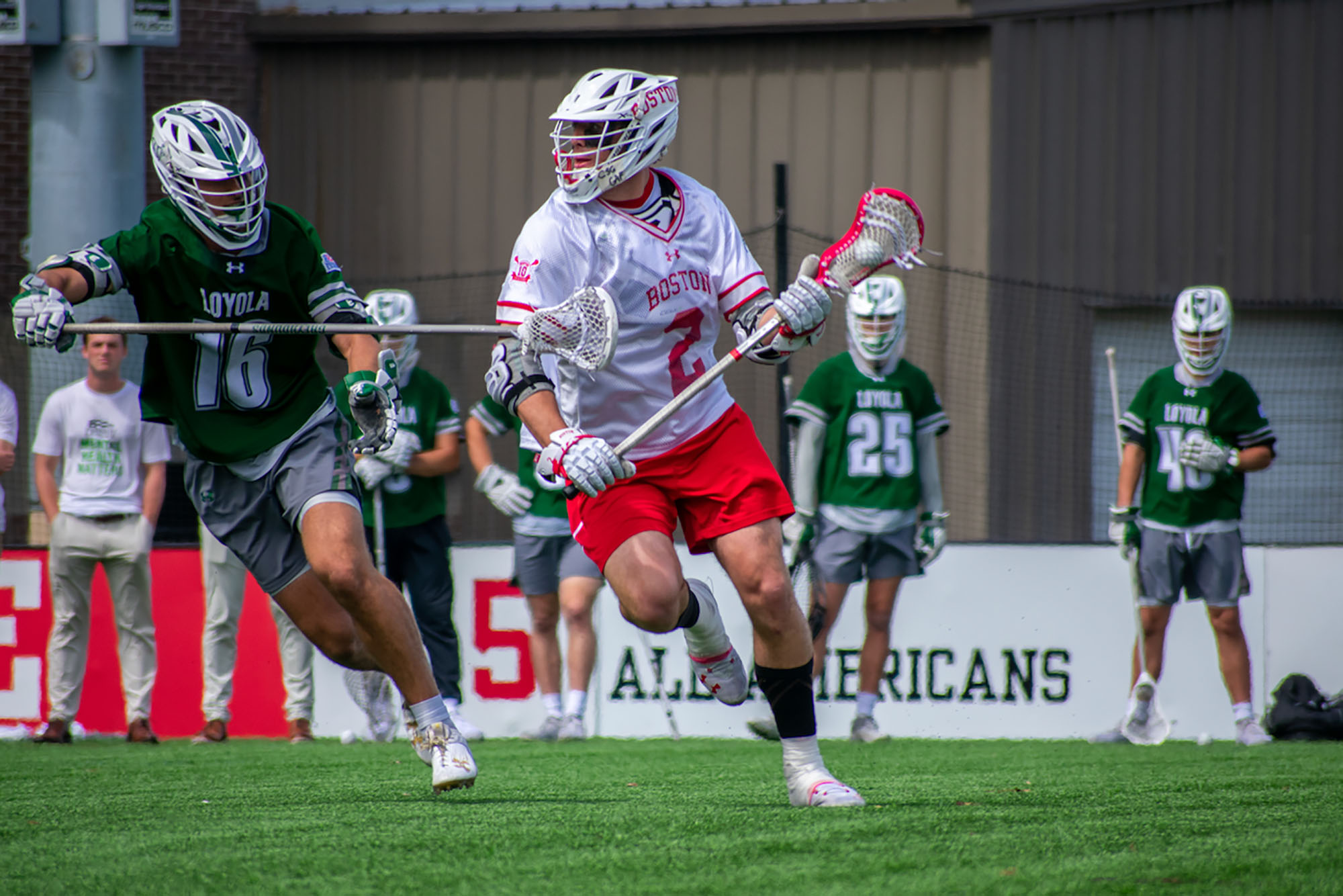 Photo: A BU lacrosse player swerves around the opposing team on a the lacrosse field. The BU player wears his white uniform with red detailing and the opposing team member wears a green uniform with white detailing.