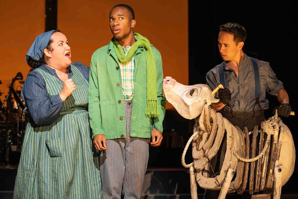 Photo: A group of 3 actors on stage, one wearing a dress, another a suit, and the third a cow costume