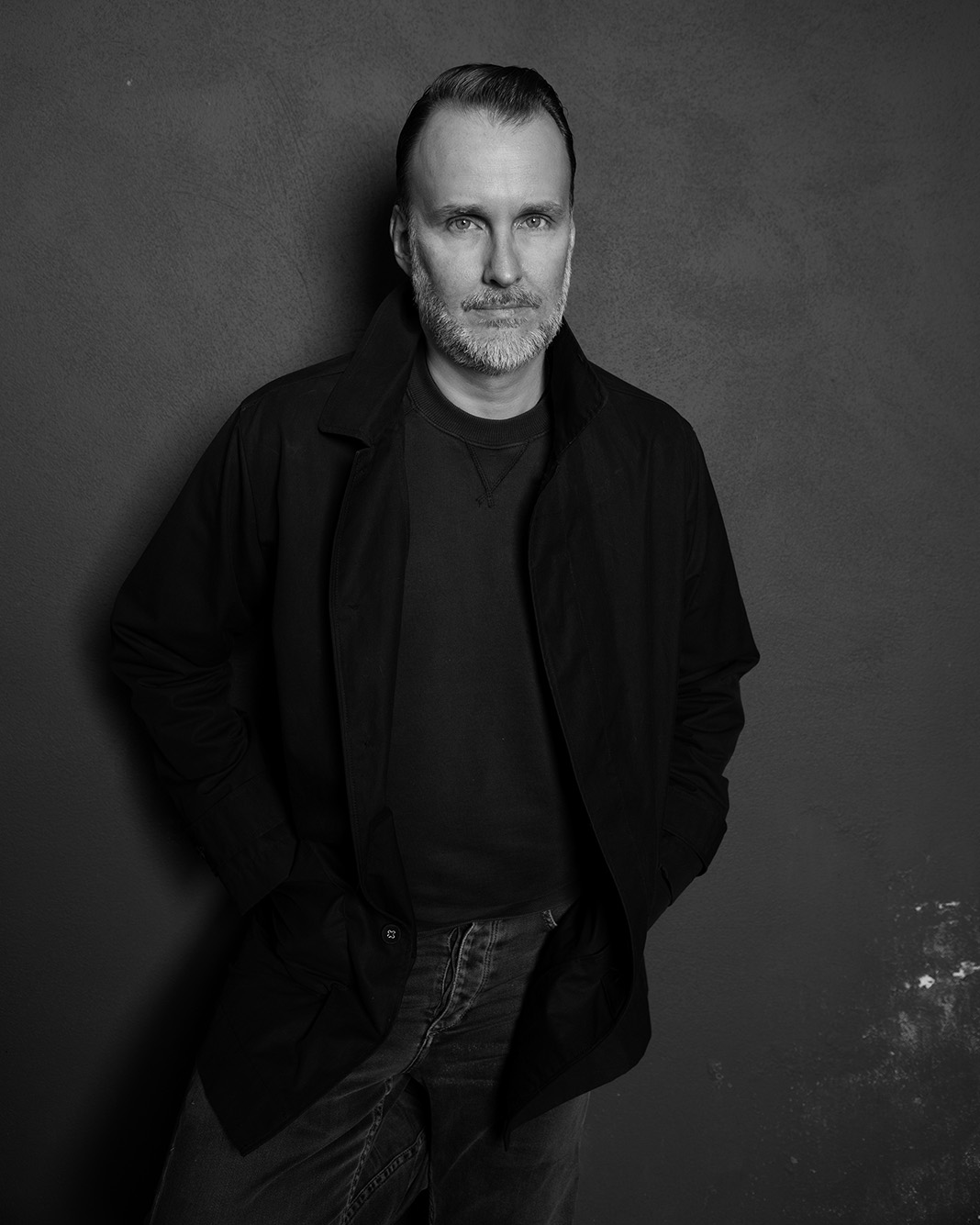 Black and white Photo: Stephen Kijak, a white man wearing a dark shirt, black jacket, and jeans, poses in front of a blank wall.