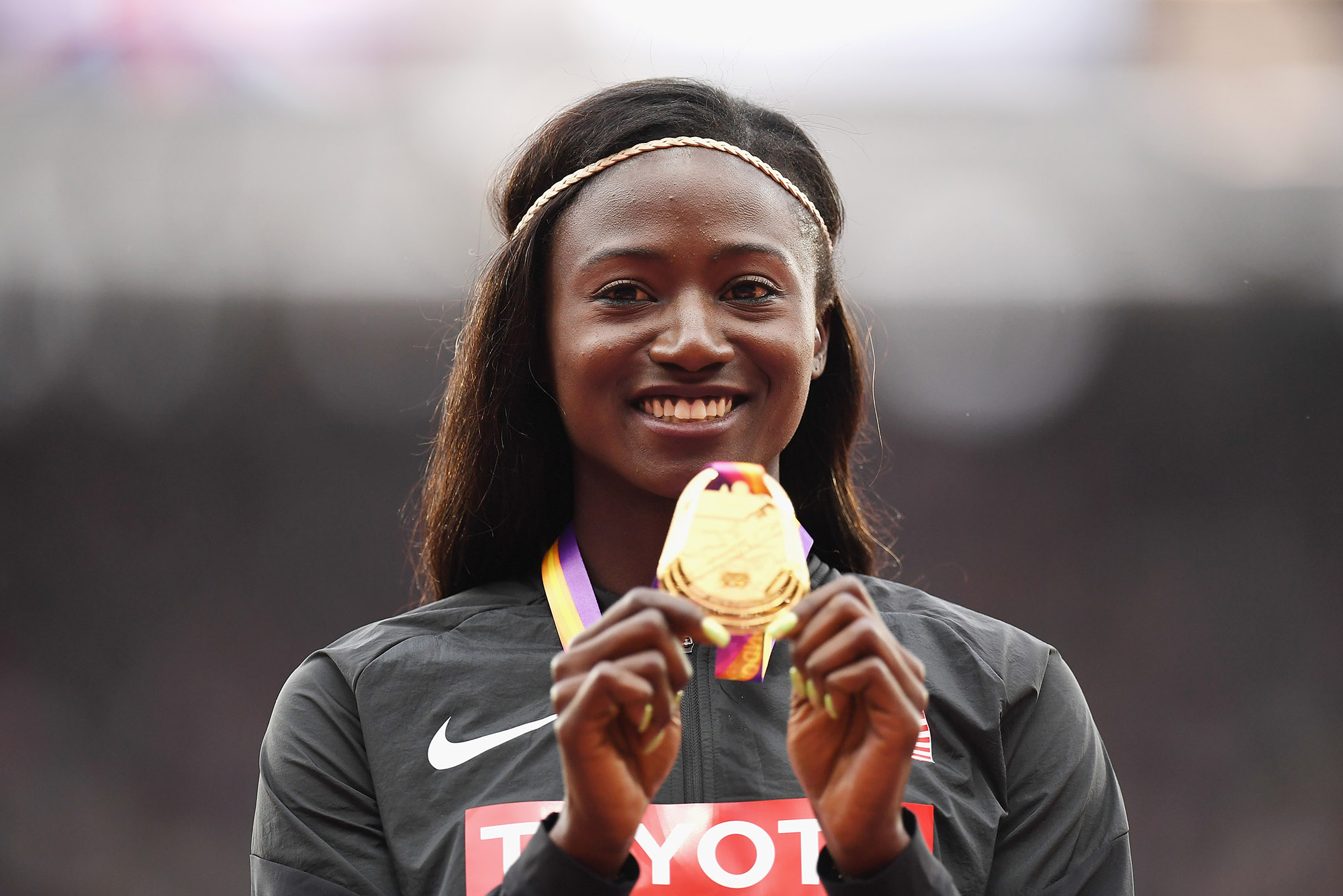Photo: Tori Bowie, A Black woman with straight, dark brown ahir and wearing a black Nike track suit, holds up a gold World Athletics Championship medal.