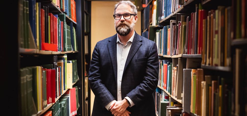 Photo: Mark Newton, a white man with dark and light gray hair, beard, and mustache and wearing glasses, a light blue collared shirt, and navy blue blazer and pants, poses with hands clasped in front of him between two large large library shelves filled with books.