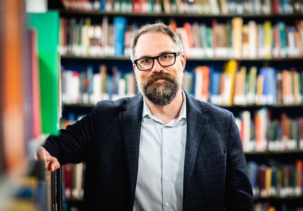 Photo: Mark Newton, a white man with dark and light gray hair, beard, and mustache and wearing glasses, a light blue collared shirt, and navy blue blazer and pants, leans against a small bookcase and poses in front of a large library shelf filled with books.