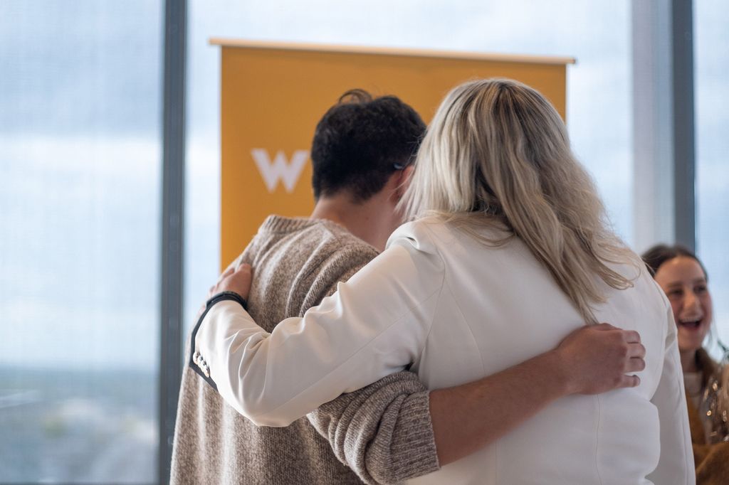 Photo: View of two people hugging. They stand in front of a yellow sign and large window as they hug with one arm over each other's shoulders.