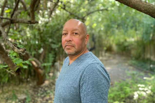 Photo: A horizontal portrait shot of a Black man, Carl Phillips, a Pulitzer Prize winner poet. He is bald and wears a thing blue-gray shirt. The setting is a wooden area.