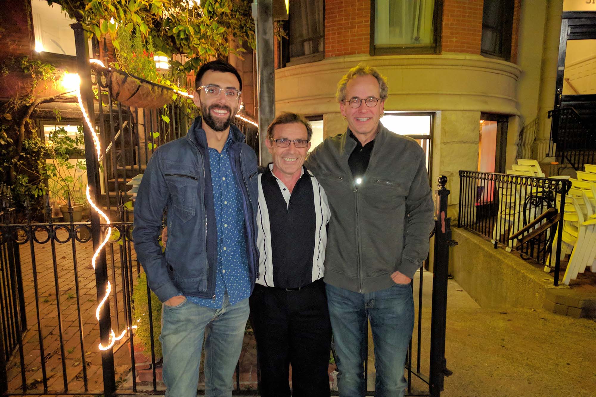 Photo: Three men stand to take a photo. It is night time and there fairy lights behind the one that stands on the left. From the left, a taller man with dark hair and a beard wears a denim button-up and a denim jacket. In the middle, a shorter man with dark hair and a black and white button-up, and lastly, on the right, a taller man with graying hair, a black t-shirt, and a gray sweater. They all wear glasses and are smiling for the photo.