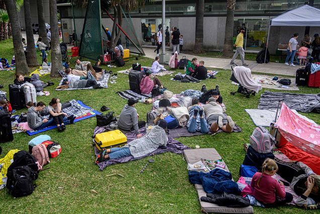Photo of Ukrainian refugees awaiting entry into the US. They sit and lay on blankets on a patch of grass. Many wearing sweatshirts and jeans and look away from the camera towards a gray tent and wall. The refugees sit amongst their suitcases and there is a mix of families and groups of young people.
