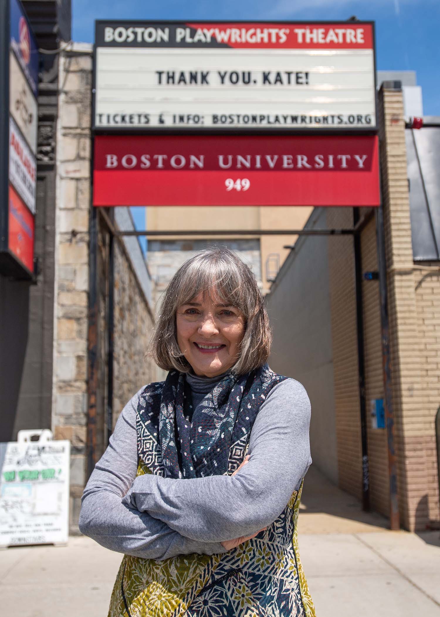 Photo of Kate Snodgrass, a middle aged White woman with a gray bob, who wears a dark blue scarf with white pattern. She stands, smiling, with her arms across on the sidewalk in front of the Boston Playwright's Theater entrance, with a red BU sign at the bottom. The marquee sign at the top reads "thank you Kate!"