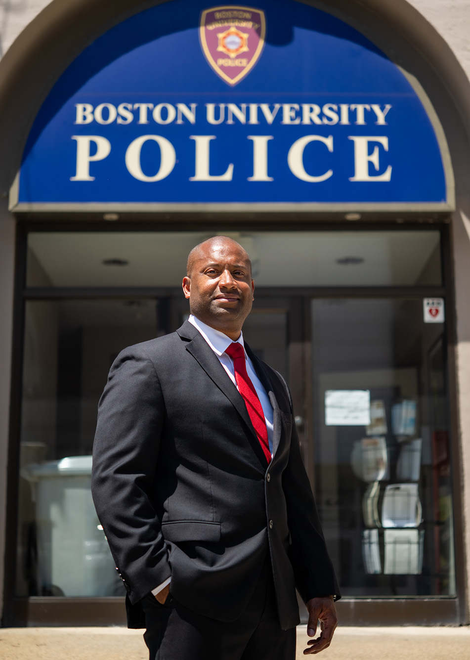 Photo: Robert Lowe, a Black man wearing a black suit, white collared shirt, and red ties, poses with one hand in pocket in front of the Boston University Police station. Behind him, the station can be sign with large blue sign that reads "Boston University Police".