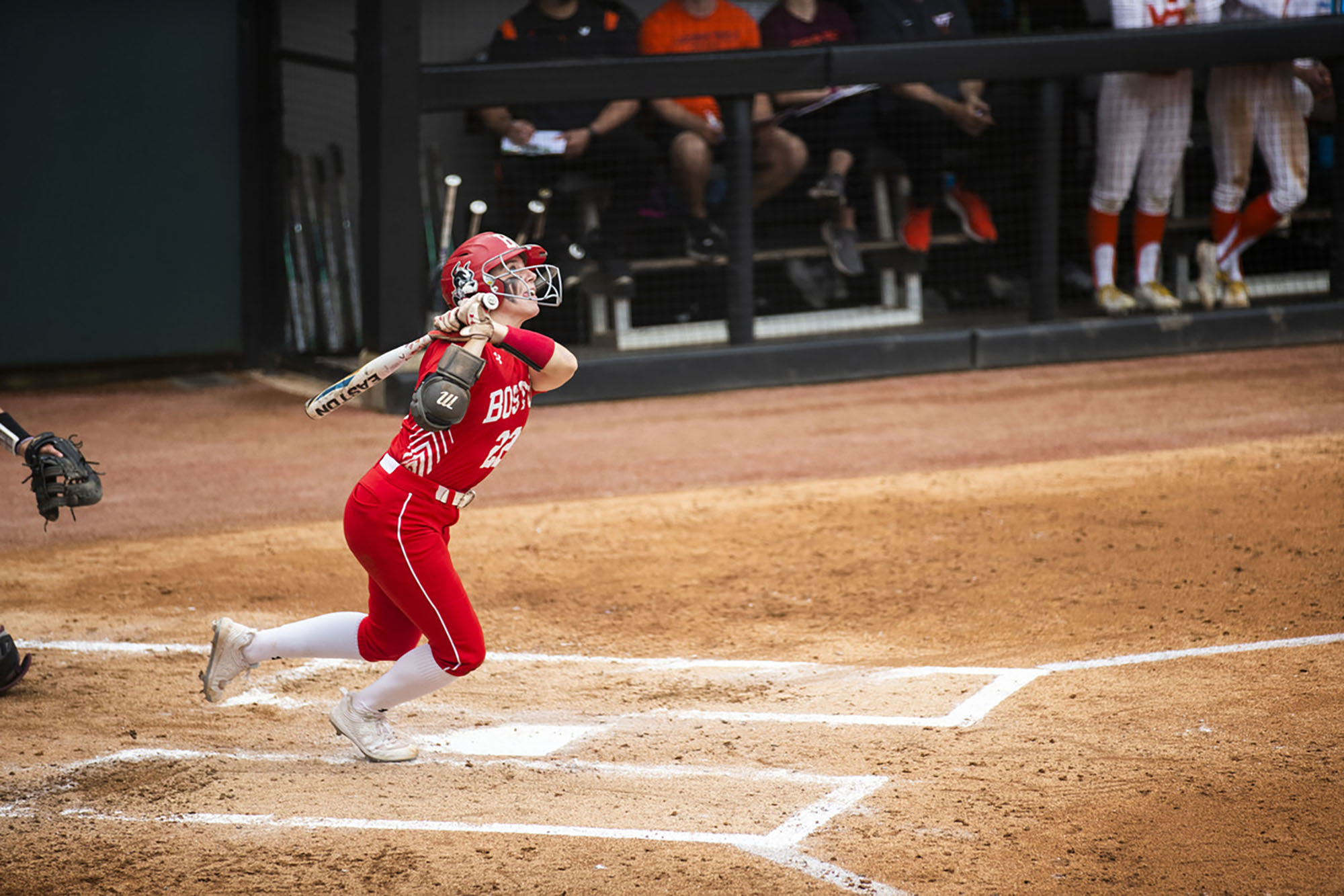 Photo: Caitlin Coker, a young woman wearing a red Bu softball uniform and helmet, holds a softball bat and watches a flyball sail in this year’s NCAA Tournament Athens Regional. She stands at home base as she looks up.