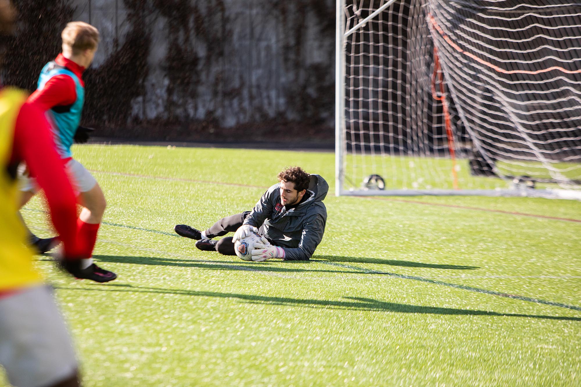 Photo: Action shot of Francesco Montali as he stands and raises his hand in a soccer goal. A young white man with curly brown hair and wearing a grey jacket and black short and spandex, diving and catching a soccer ball before it rolls into the goal. A player wearing a red jacket, white shorts, and blue vest runs in front of him.