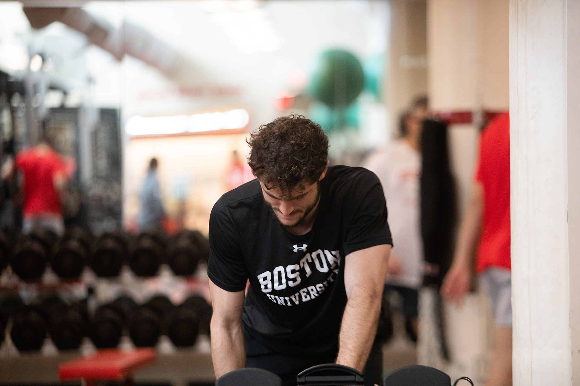 Photo: Action shot of Francesco Montali as he stands and raises his hand in a soccer goal. A young white man with curly brown hair and wearing a black shirt that reads "Boston University" in white letters, bends over slightly and lifts a large dumbbell with his left arm. A gym with various people are shown in the blurred background.