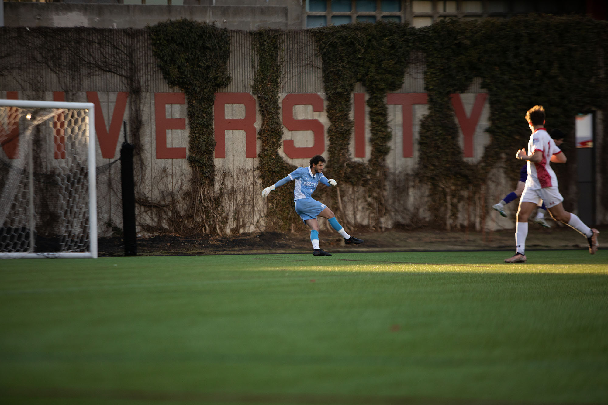 Photo: Action shot of Francesco Montali as he stands and raises his hand in a soccer goal. A young white man with curly brown hair and wearing a light blue and white soccer uniform and white gloves, is shown mid-kick as he sends a soccer bowl down Nickerson soccer field.