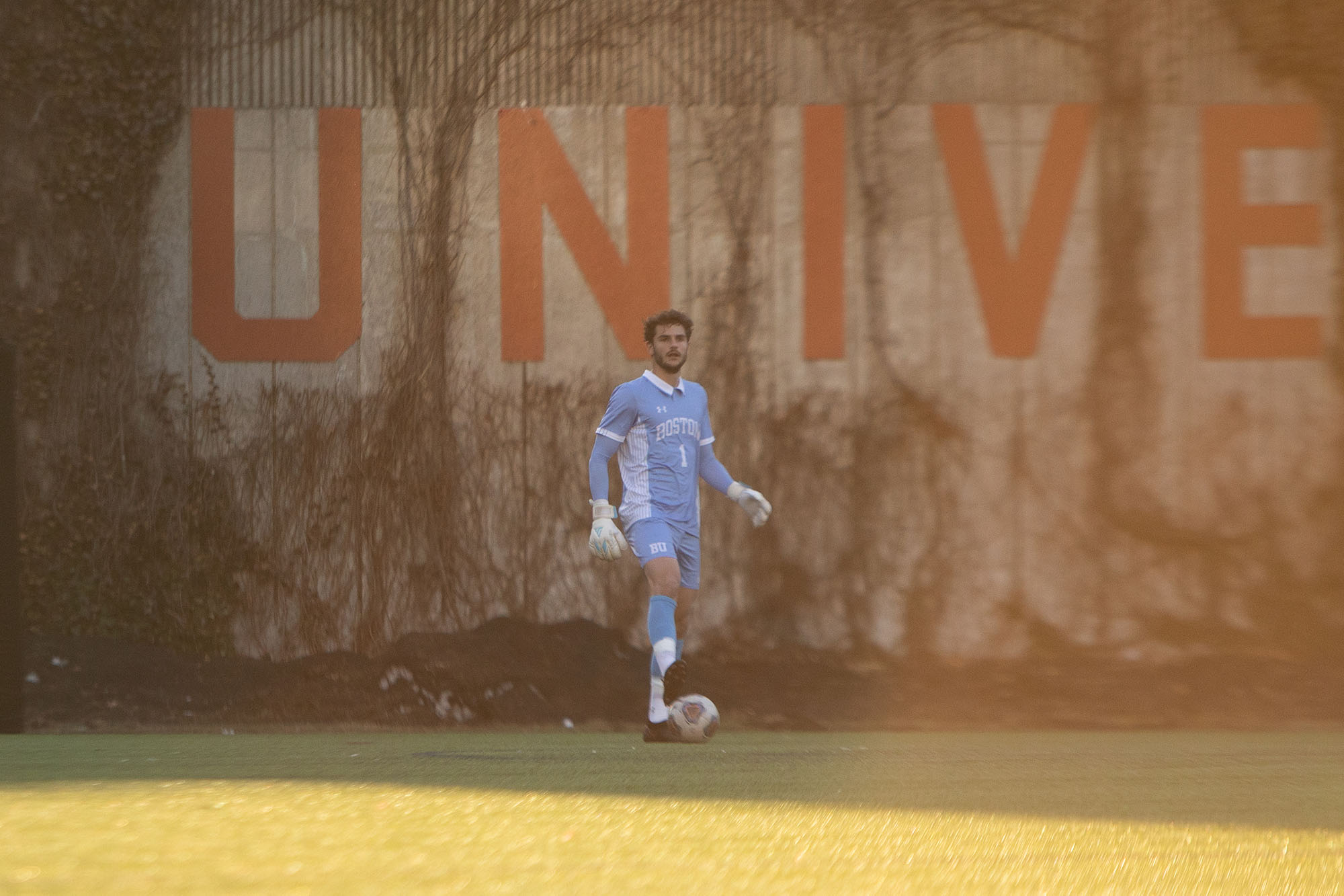 Photo: Action shot of Francesco Montali as he stands and raises his hand in a soccer goal. A young white man with curly brown hair and wearing a light blue and white soccer uniform and white gloves, stands with one foot on a soccer ball as he looks towards the field. He stands in front of vine-y wall with large red letters that read "university".