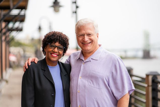 Photo: A black woman and white man stand side by side with smiles on their faces in front of a body of water on an overcast day