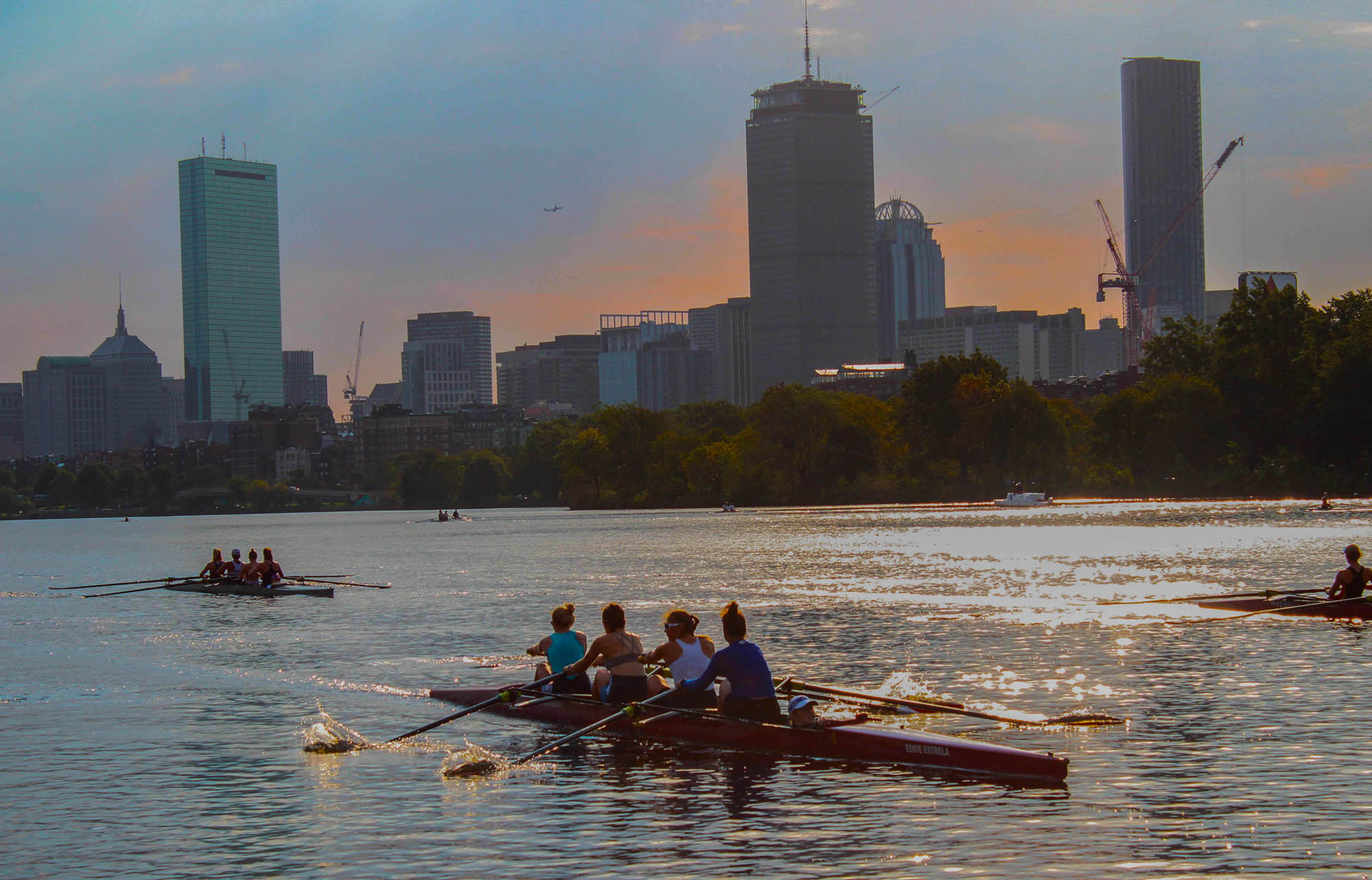Photo: A boat full of women rowers are shown rowing down the Charles River in Boston as the sun sets. Cit buildings can be seen in the distance as they row along with several other rowers on the water.