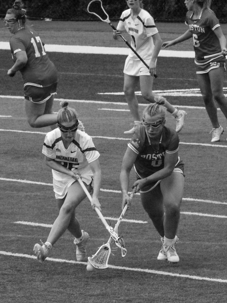 Photo: Black and white action shot of Ellen Monahan as she plays lacrosse. A young white woman wearing a BU lacrosse uniform and goggles battles for the ball against an opposing athlete with her lacrosse stick. Other athletes on both teams can be seen moving around the field behind them.