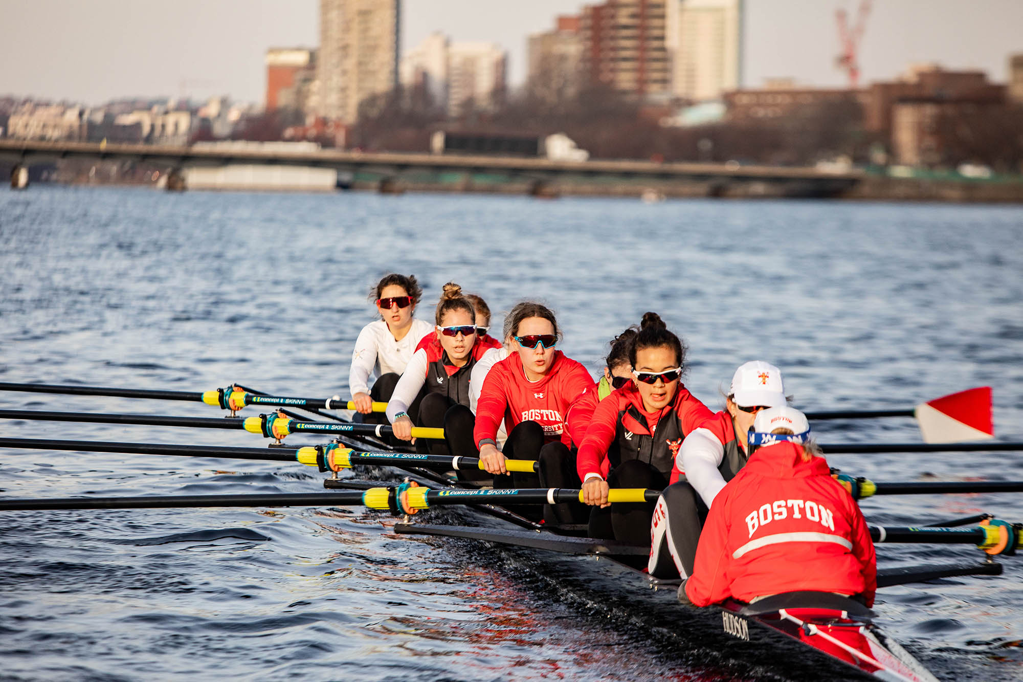 Photo: A boat full of women rowers are shown rowing down the Charles River in Boston on a gloomy day. The rowers wear BU gear and sunglasses as a city skyline can be seen in the blurred background.