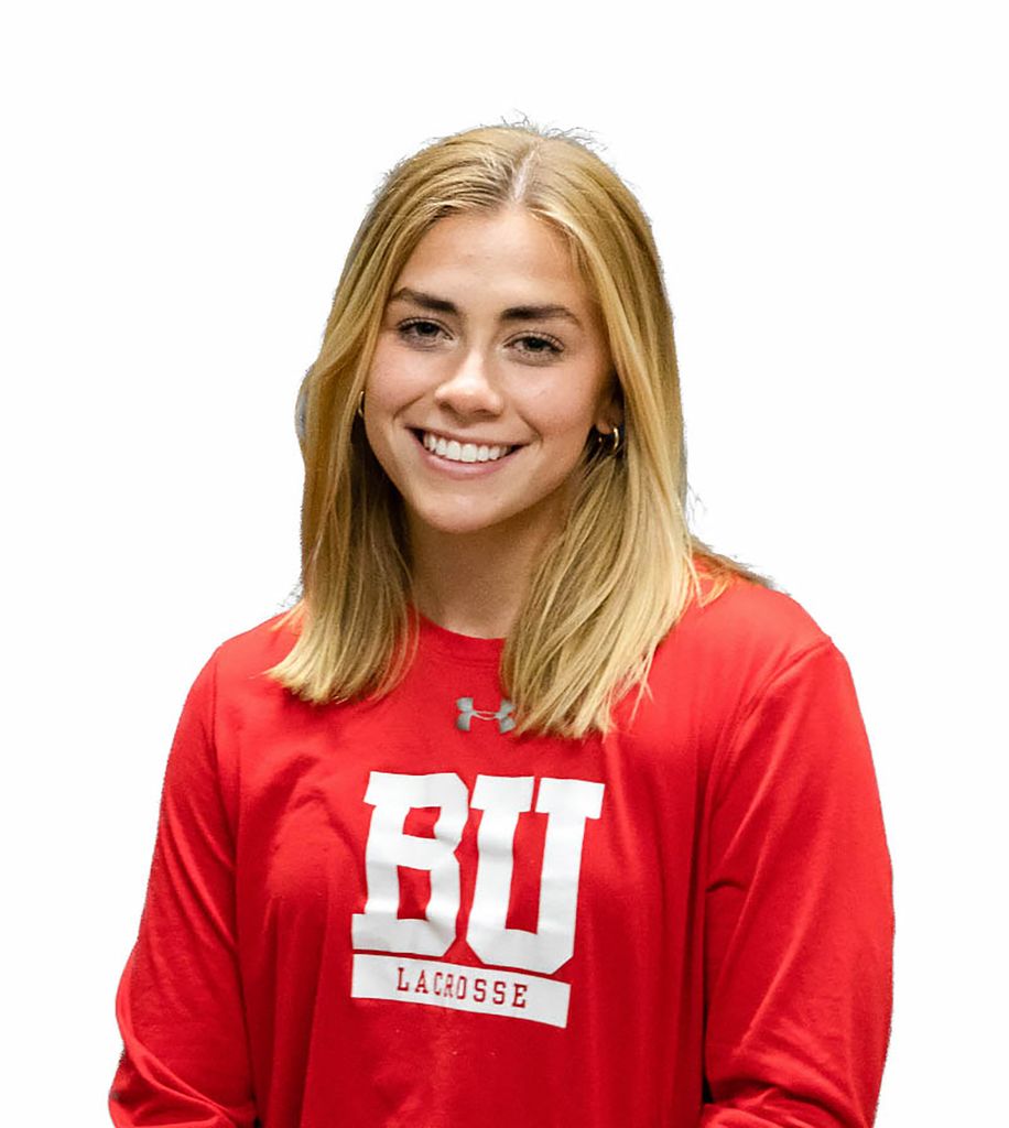 Photo: Headshot of Ellen Monahan. A young white woman with shoulder length blonde hair and wearing a red, BU lacrosse long sleeve shirt poses in front of a white background.