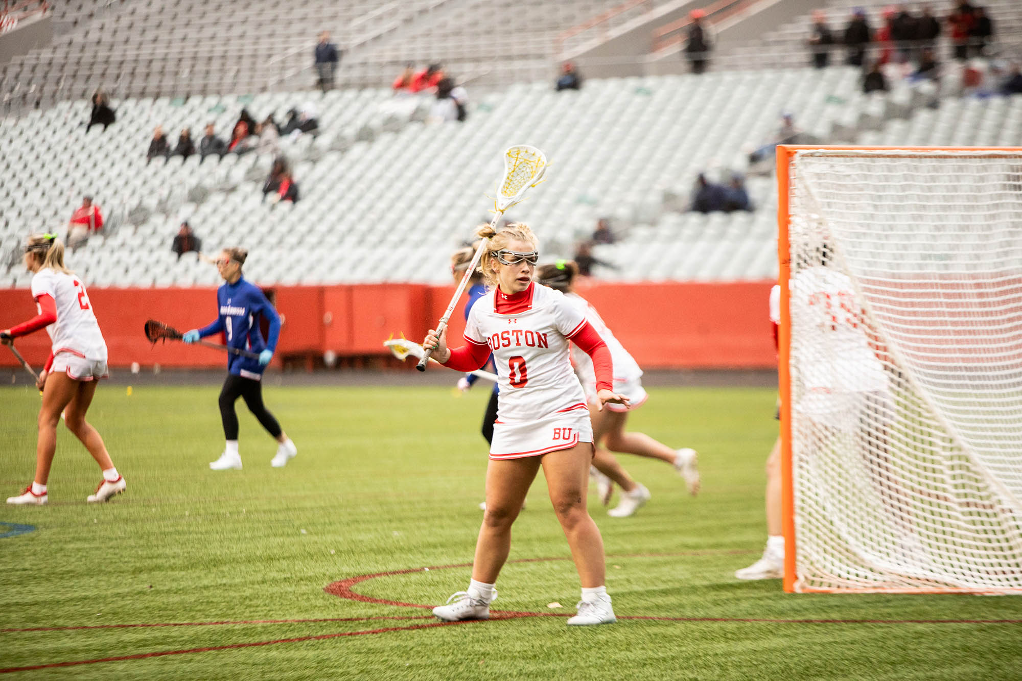 Photo: Action shot of Ellen Monahan as she plays lacrosse. A young white woman wearing a BU white and red lacrosse uniform and goggles looks to the right as she raises her lacrosse stick in her left hand. Other BU players and players wearing blue uniforms can be seen running on the field around her.