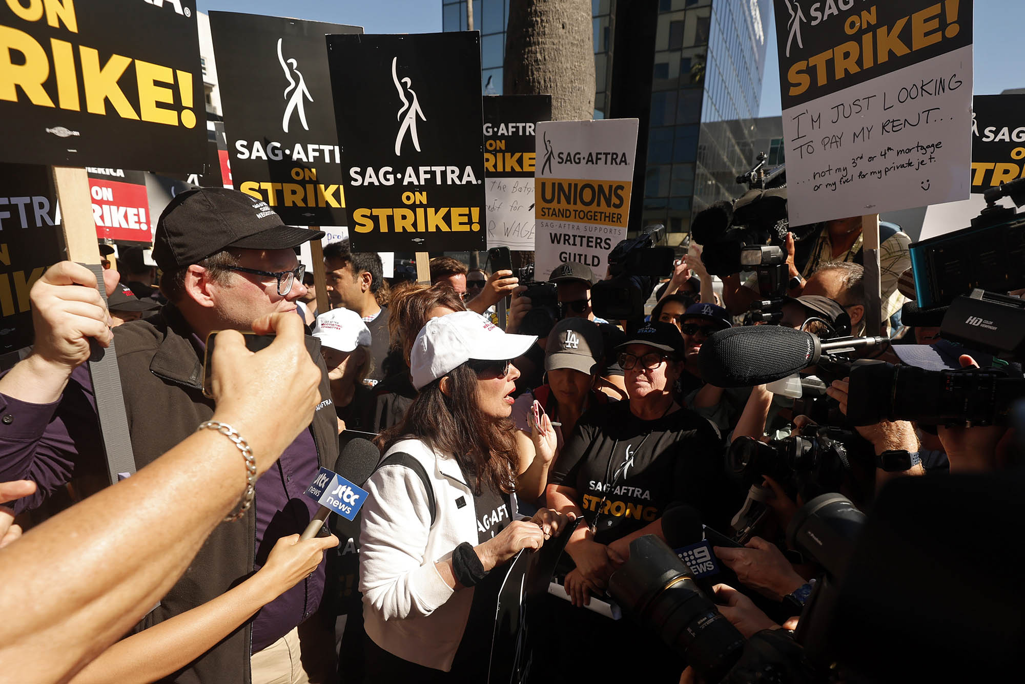 Photo: Fran Drescher, President of SAG, is swarmed by news outlets as she heads to a press call. She is wearing all white and stands in the middle while cameras and microphones surround her.