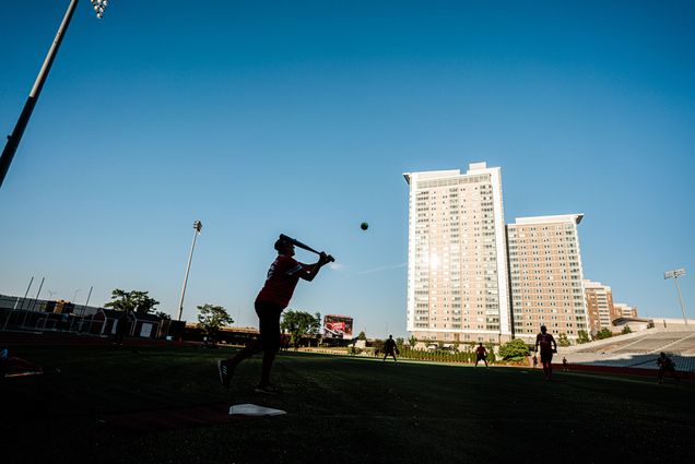Photo: A batter swings to hit a ball on the field with buildings in the background.