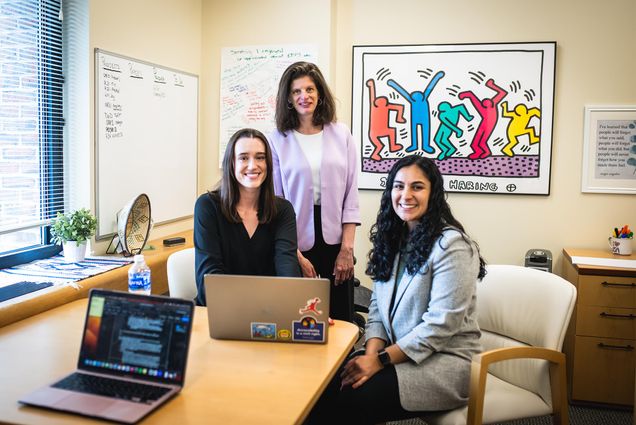 Photo: Three women gather around a desk, 2 sitting and one standing. All are looking towards the camera and smiling. There is a laptop on the desk and a painting in the background.