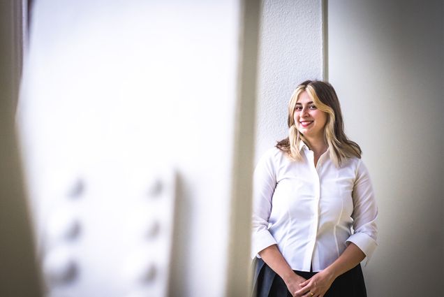 Photo: A woman in a white collared shirt stands with hands clasped and a soft smile in front of a white wall.