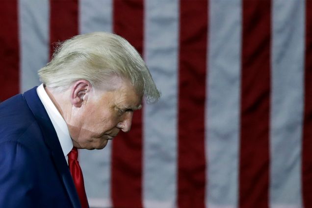 Photo: Donald Trump hangs his head while wearing a suit. Background is an american flag