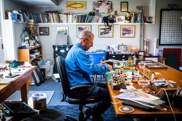 Photo: Scott Biron makes a fly at his New London, NH home. An older white man wearing glasses, a long-sleeved blue collared shirt, and jeans sits at a crowded desk and works on a fly. Fly tying materials and paraphernalia can be seen around the office.