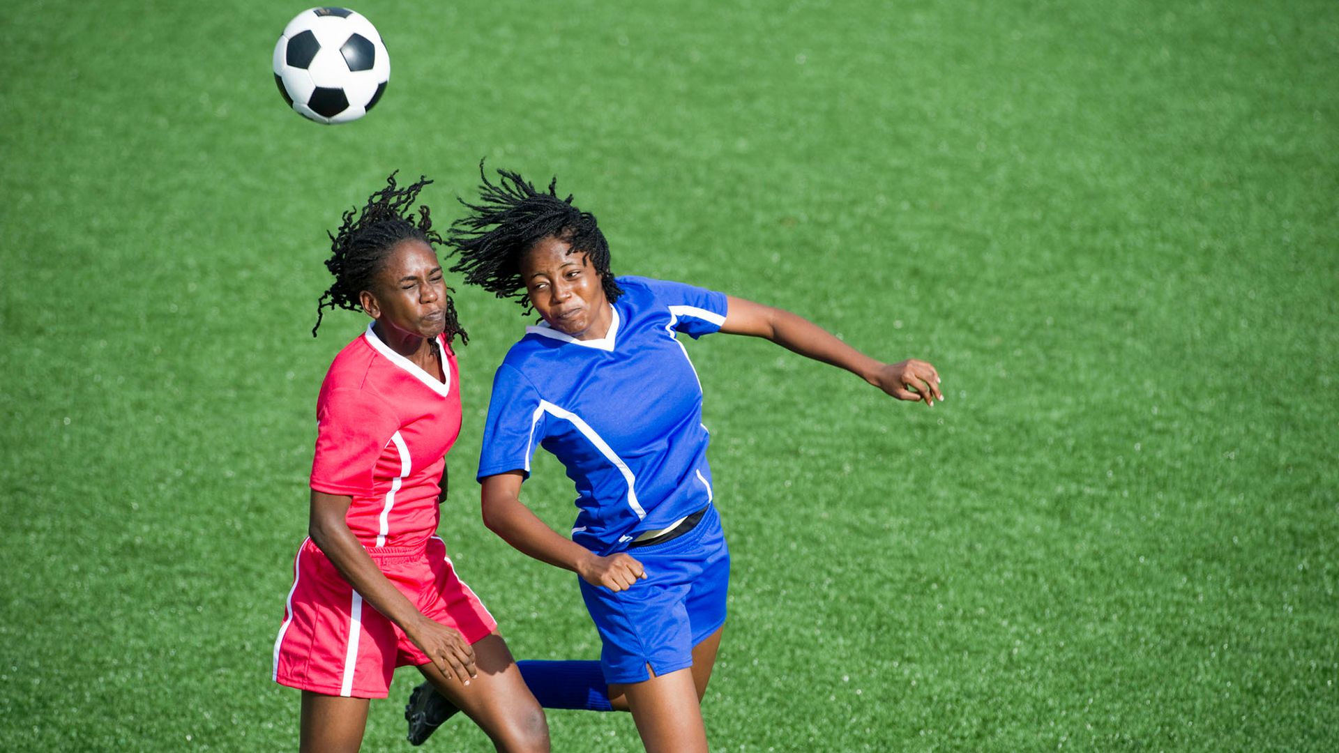 Photo: Two female soccer player, one in a red uniform and the other in blue, jump to head butt a ball flying through the air.