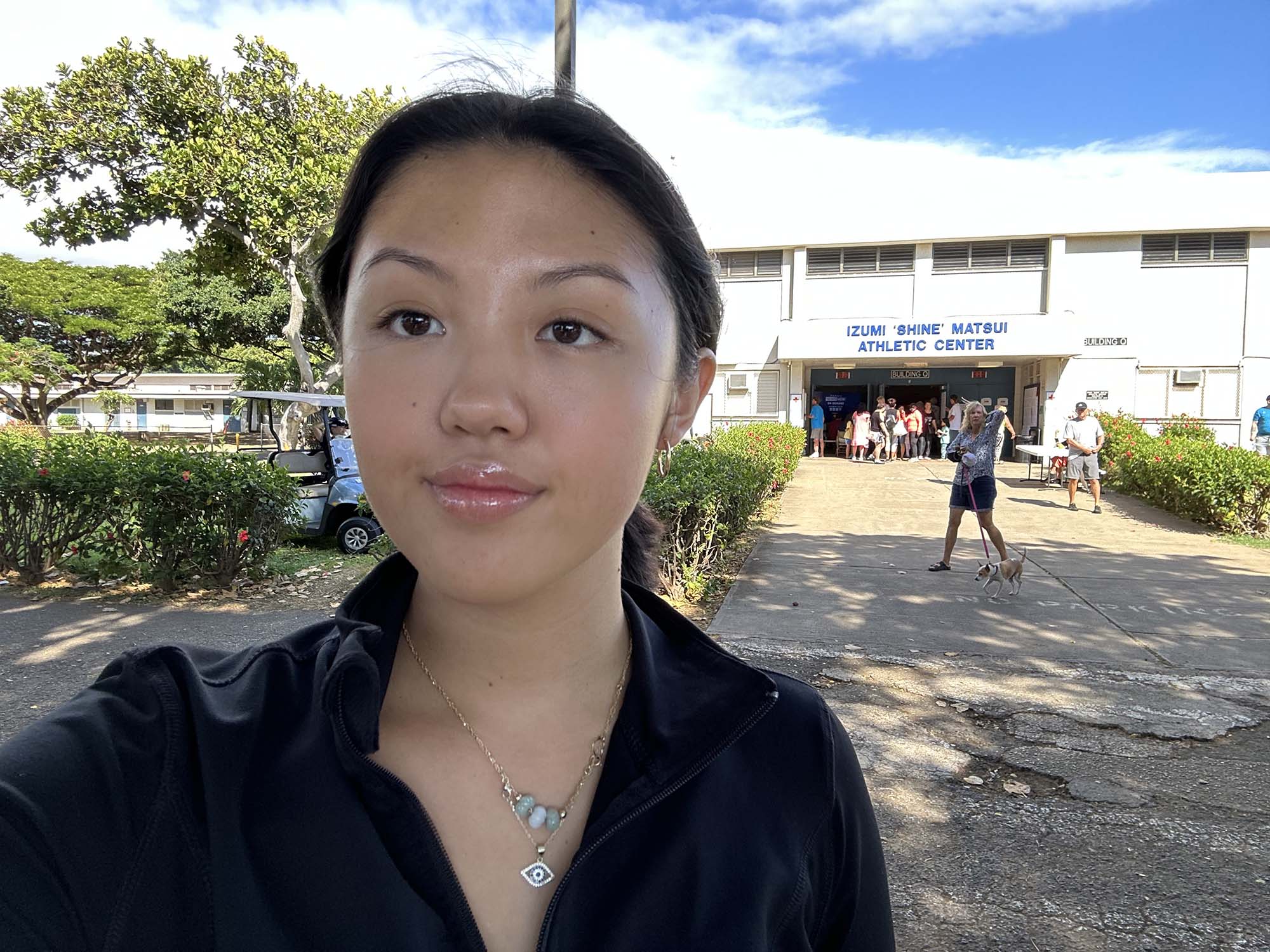 Photo: Noelle Lo, a young Hawaiian woman wearing a black collared shirt takes a selfie in front of a high school building on a sunny day.