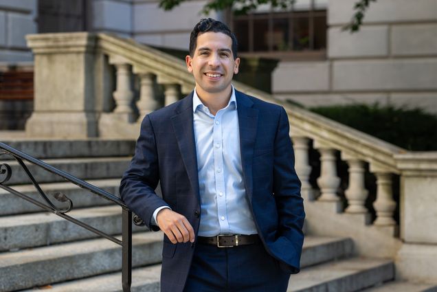 Photo: A man in a navy suit with short dark hair smiles on the steps of the Massachusetts State House, a large ornate government building.