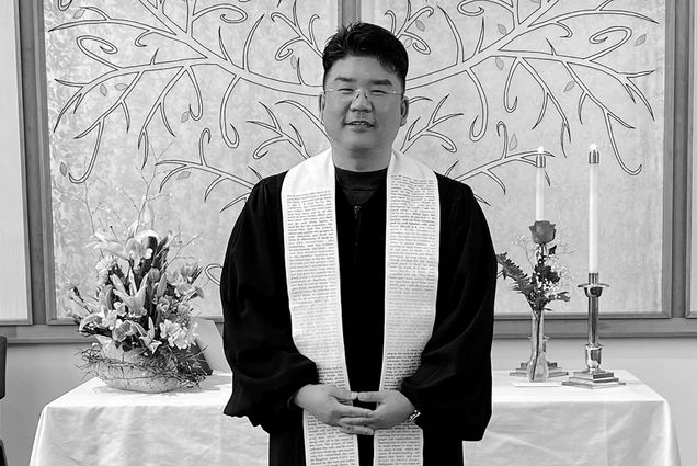 Photo: A Korean man, Jinwoo Chun, clasps his hands in front as he stands to pose for the photo. He wears his religious robes in front of a table with a white tablecloth in a cross on the background.