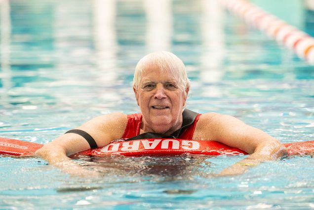 Photo: Photo: Tom Gilmore, professor of Biology and director of the graduate program in Molecular Biology, Cell Biology & Biochemistry, a white man with grey hair, holds onto a lifeguard floatation spear as he smiles for a photo in the FitRec pool.